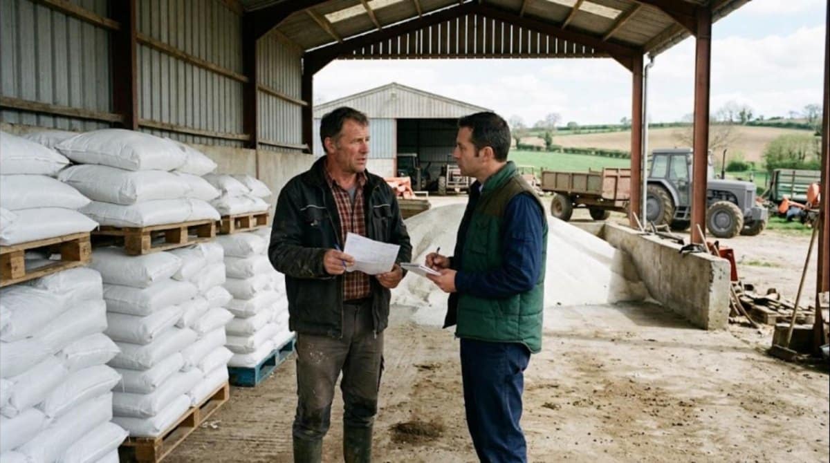Éleveur et vendeur discutant d'une facture devant des sacs d'engrais azoté sous un hangar de négoce agricole